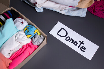 'Donate' sign handwritten with black letters. A box with clothes and a pile of clothes nearby on a grey table.clothes donation concept. copy space. High quality photo