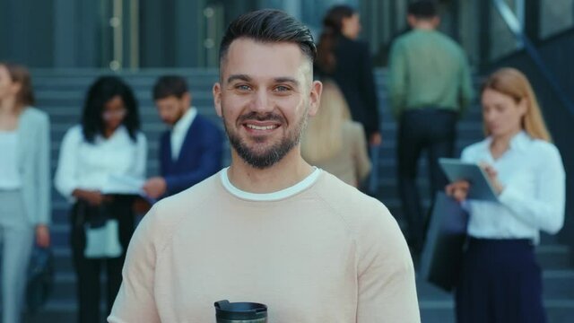 Outdoor portrait of young bearded businessman with coffee cip smiling standing outside modern corporate building department. Successful people. Business.