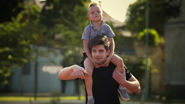 Young dad in sportswear is smiling and carrying tired kid boy on his shoulders, showing something to him while walking along the stadium. Slow motion