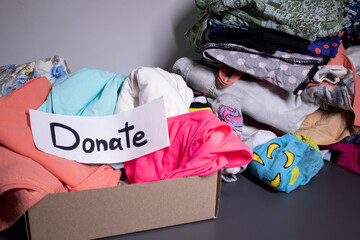 'Donate' sign handwritten with black letters. A box with clothes and a pile of clothes nearby on a grey table.clothes donation concept. copy space. High quality photo
