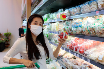 Asian woman wearing protective face mask push and hold shopping cart in supermarket department store. Girl, looking grocery to buy  some food. New normal after covid-19. Family concept.
