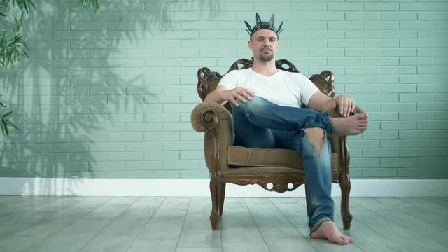 barefoot man in a t-shirt and jeans in a leather crown sits in an armchair