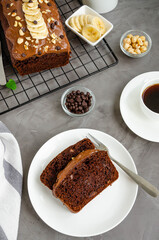 Homemade chocolate banana bread with chocolate cream, banana slices and nuts on top on a dark concrete background. Vertical orientation. Top view.
