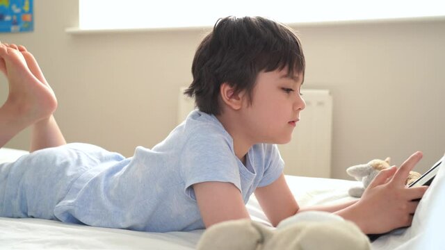 6-7 year old boy playing with dog toy and watching cartoon on tablet,Happy child lying in bed playing game in the morning, Cute Kid having fun and relaxing on his own in bed room,New normal lifestyle 