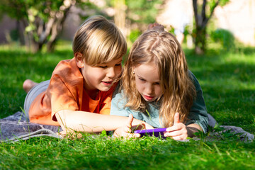 Young caucasian boy and girl use a mobile phone and smile while lying on the grass. Sunny summer day in a park