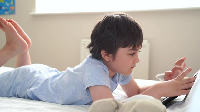 6-7 year old boy playing with dog toy and watching cartoon on tablet,Happy child lying in bed playing game in the morning, Cute Kid having fun and relaxing on his own in bed room,New normal lifestyle 