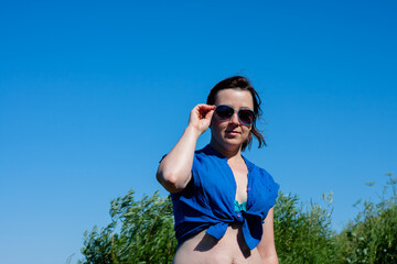 a young sexy woman in a swimsuit and a blue shirt adjusts her sunglasses. in summer
