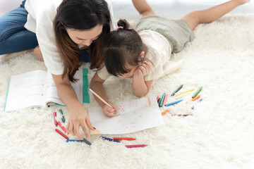 top view of happiness Asian mother teaching her little daughter to draw by pencil and crayon, lie on the floor of the house, family joyful together in the house on holiday.