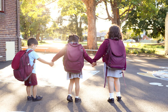 Three school children with backpacks on, walking through playground holding hands.