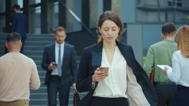 Appealing young caucasian businesswoman with smartphone walks in corporate neighbourhood outdoors. Business people lifestyle. Social distancin Technology.