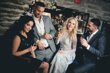 Party, holiday with friends concept. Four people with champagne glasses celebrating and toasting in restaurant. Two men and two women in elegant evening clothes, suits and dresses night out indoors