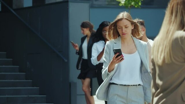 Young stylish businesswoman using smartphone walking nearby corporate building with her hair blowing in the wind. Office people. Communication. Technology. Lifestyle.