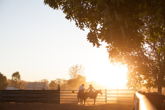 Two horse riders meet at stockyard gate at sunset