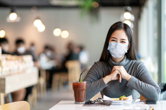 Young asian lady smiling with eyes under medical mask feeling safety relax at restaurant or canteen after coronavirus pandemic opening business eat lunch for breakfast with social distancing concept.