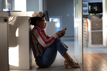  afro brazilian business woman with mobile sitting on the floor alone in office
