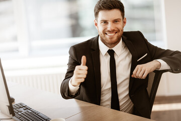 Business concept. Successful young businessman at work. Manager sitting at the office table happy showing thumb up. Man smiling in suit indoors on glass window background