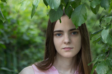 A young girl with long blonde hair stands under a tree and hides part of her face behind leaves and branches.