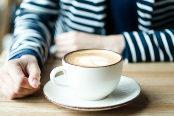 A cup of cappuccino on the wooden table and a woman in a blu jacket with white lines. Beige, blue, coffee, cappuccino, woman.