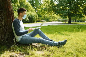 Handsome young man in medical mask works on a laptop under a tree in a green park