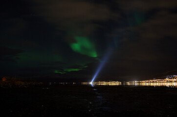 aurora borealis over a boat searchlight on the fjord reflecting of the surface with illuminated settlement in the background