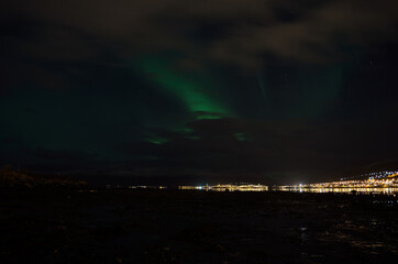 Naklejka na ściany i meble aurora borealis, northern lights over mountain landscape with new snow reflecting on beach sand and fjord water illuminated by the whale island settlement lights in late autumn