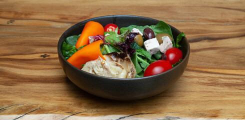 Fresh salad with greens, olives, tomatoes, grilled artichoke, peppers and feta cheese, on a sycamore wood table with a natural flame pattern.