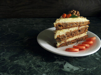A slice of carrot cake on a white saucer, decorated with walnuts, dried sea buckthorn and green leaves. Background - green marble countertop.