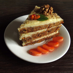 A slice of carrot cake on a white saucer, decorated with walnuts, dried sea buckthorn and green leaves. Carrot lies nearby, the background is a green marble countertop.
