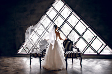 The bride in a white wedding dress stands near two striped armchairs, against the background of a triangular design window