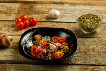 On a wooden table is a black dish of boiled mung bean with mushrooms and vegetables.