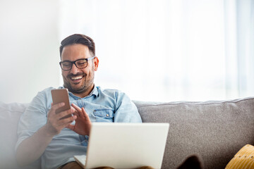 Businessman working at home using lap top. Home office. Young man sitting relaxed on sofa with laptop and smartphone. Happy young man using a mobile phone on the sofa at home
