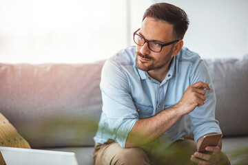 Smiling casual freelancer using smart phone and laptop while sitting on the sofa. Smart apps make for smarter financial planning. 