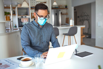 A young man fulfills recommendations to prevent the spread of the virus in the office. He is putting on a protective medical mask in front of a computer.