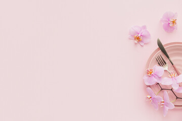 Flower and table settings overhead composition on light pink background. Pink ceramic plates, cutlery and perple orchid flowers. Holiday modern table decoration. Romantic date.