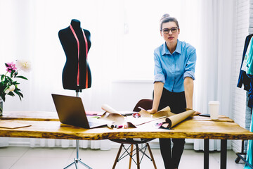 Full length portrait of attractive design owner of fashion atelier in optical spectacles looking at camera standing at desktop with sketches and sewing instruments for tailoring stylish clothes