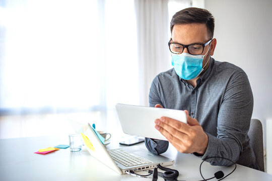 Man surfing the internet wearing a facemask. Businessman looking at a tablet PC wearing a healthcare face mask. A young man in a medical mask works in the office during an epidemic of coronavirus