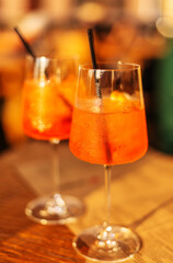 Two glasses of wine with aperol spritz stand on a wooden table in a cafe, in the rays of the setting sun. Italian alcoholic drink with oranges and ice. Close-up, selective focus, vertical composition.