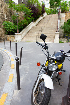 Close up image of a yellow white vintage motor bike parked by the street. On the blurred background, there are stone stairs trees and street lamps creating a scenic city view.