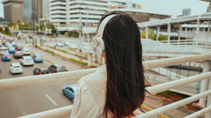 Fototapeta na wymiar Asian girl stands on a bridge over road traffic and in white headphones listens to music.