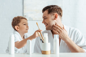 selective focus of smiling man touching face while adorable son taking toothbrush near toiletries