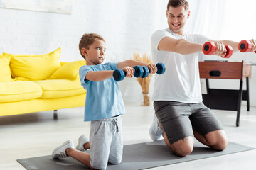 Fototapeta na wymiar smiling father with son exercising with dumbells while standing on knees on fitness mat