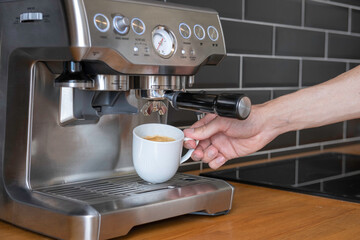 A man makes coffee at home in a gray carob metal coffee machine (coffee maker) and shows ready coffee in a white cup against a background of black tile wall and wooden tabletop