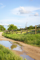 Obraz premium Forststraße mit einer Wasserpfütze und einem Weidezaun blauer Himmel und Wolken