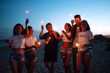 Group of young people having fun with sparklers outdoors at the sea shore. People celebrating new year's day at the beach. Beach party, rest, vacation.