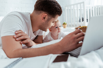 selective focus of young man using laptop while lying on bed near adorable son