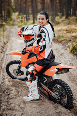 Happy brunette girl wearing motorcycle outfit sitting on her bike off-road in the woods