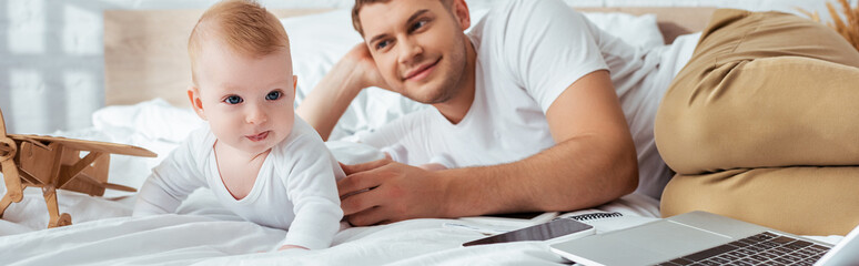 panoramic orientation of happy father and cute infant on bed near smartphone at toy plane
