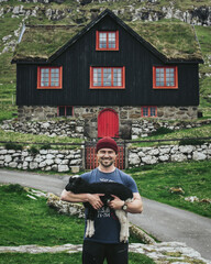 Happy man holding little sheep. Guy on the background of a house with a grass roof in the Faroe Islands. Tourism is an active life and adventure in Scandinavia.