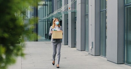 Caucasian businesswoman in medical mask walking outdoors with box of stuff leaving business. Female office worker lost her job. Unemployment rate growing at pandemic. Fired woman coming back home.