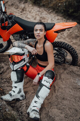 Tattooed female racer wearing motocross outfit with semi naked torso sitting on sand next to her...